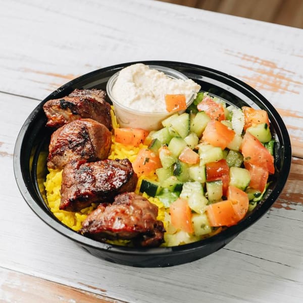 A black bowl filled with yellow rice, grilled meat, diced cucumber and tomato salad, and hummus.