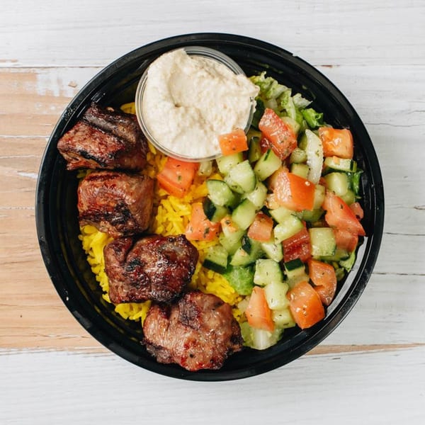 Grilled beef chunks, yellow rice, cucumber and tomato salad, and hummus in a black bowl.