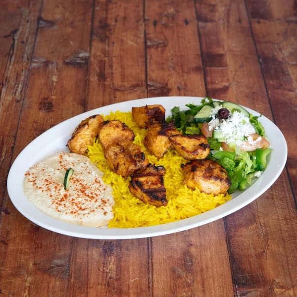Grilled chicken skewers with yellow rice, hummus, and a Greek salad on a white plate.