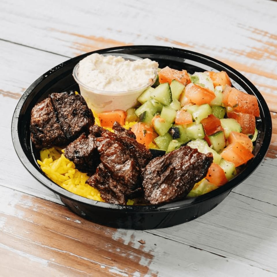 A black bowl filled with yellow rice, grilled meat, a side of chopped salad, and a small cup of sauce.